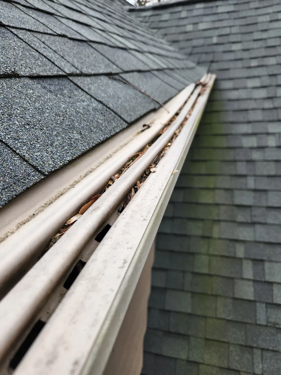 Weathered white plastic gutter guards installed along a roofline in Alamance County NC, panel slots packed with debris and surfaces stained from years of sun and rain